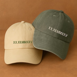 May include: Two baseball caps, one tan and one olive green, are displayed. Each cap has the Roman numeral date "XX.XII.MMXXV" embroidered on the front. The caps are made of a soft fabric and have a classic, casual style.
