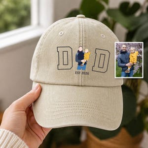 May include: A beige baseball cap featuring the text DAD with a stylized illustration of a person holding a child in the center, and the text EST 2026 below it. An inset photo shows the original image of a person holding a child in a yellow jacket.