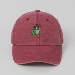 May include: A maroon baseball cap with a curved peak. The cap has an embroidered design of a green frog wearing a brown cowboy hat. The frog has white eyes and is in the centre of the cap.