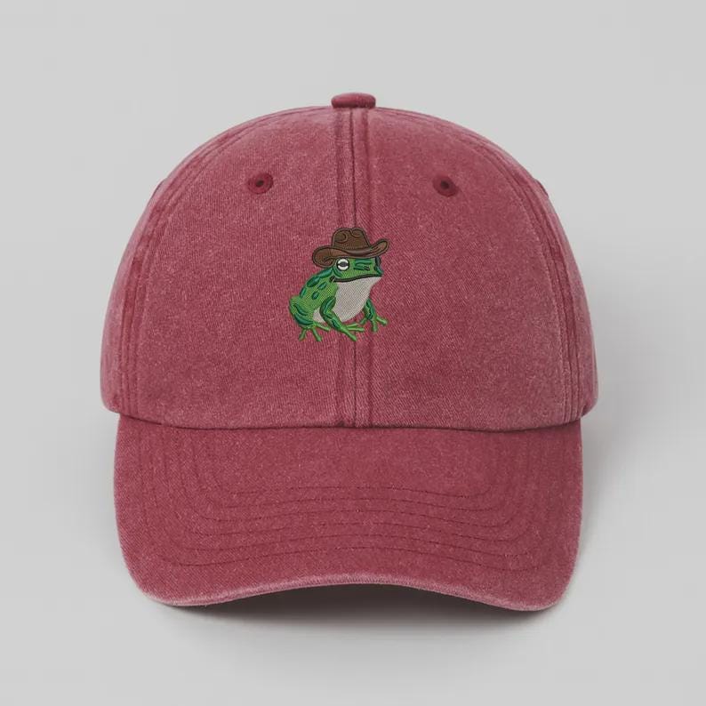 May include: A maroon baseball cap with a curved peak. The cap has an embroidered design of a green frog wearing a brown cowboy hat. The frog has white eyes and is in the centre of the cap.