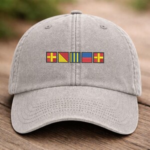 May include: A gray baseball cap with a curved brim. The cap features an embroidered design of colorful nautical signal flags across the front. The flags are red, yellow, blue, and yellow with a cross.