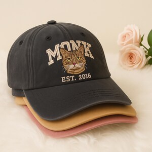 May include: A stack of baseball caps in navy, tan, and pink. The top cap is navy with the word "MONK" embroidered above a cat face and the text "EST. 2016". The caps are stacked on top of each other.