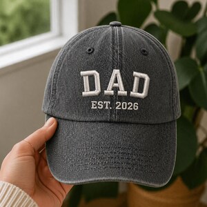 Puede incluir: Gorra de béisbol gris oscuro con la palabra "DAD" bordada en letras blancas. Debajo de la palabra "DAD" está el texto "EST. 2026". La gorra tiene una visera curva y un cierre de correa de tela.