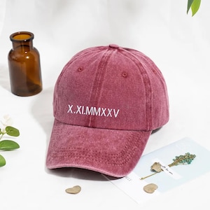 May include: A maroon baseball cap with a curved brim. The cap features the Roman numeral text "X.XI.MMXXV" in white on the front. A small brown glass bottle is in the background.