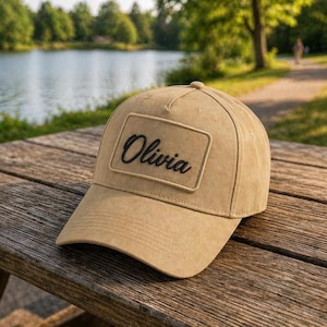 Peut inclure: Casquette de baseball beige avec le nom "Olivia" brodé en noir sur un patch rectangulaire. La casquette est posée sur une table en bois, avec un lac et des arbres en arrière-plan.