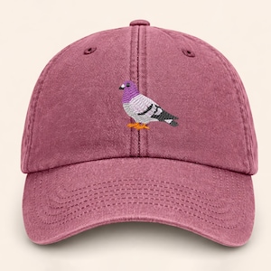 May include: A maroon baseball cap with a stitched image of a pigeon. The pigeon has a purple head, gray body, and black and white wings. The cap has a curved brim and an adjustable strap.