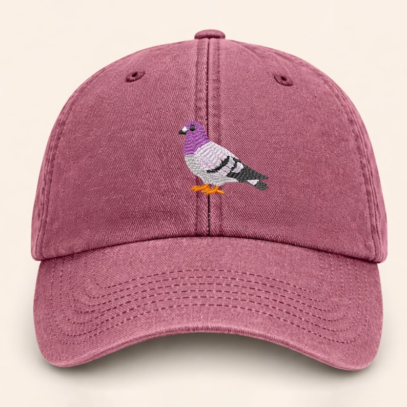 May include: A maroon baseball cap with a stitched image of a pigeon. The pigeon has a purple head, gray body, and black and white wings. The cap has a curved brim and an adjustable strap.