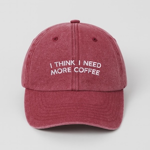 May include: A maroon baseball cap with the embroidered text "I THINK I NEED MORE COFFEE" in white. The cap has a curved peak and a fabric strap closure. The hat is made of a soft, textured material.