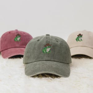 May include: Three baseball caps in red, green, and beige. Each cap has an embroidered green frog wearing a brown cowboy hat. The caps are displayed on a white, textured surface.