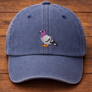 May include: A blue baseball cap with a curved brim. The cap features an embroidered pigeon design with purple, gray, black, and orange accents. The cap has a button on top and a strap in the back.
