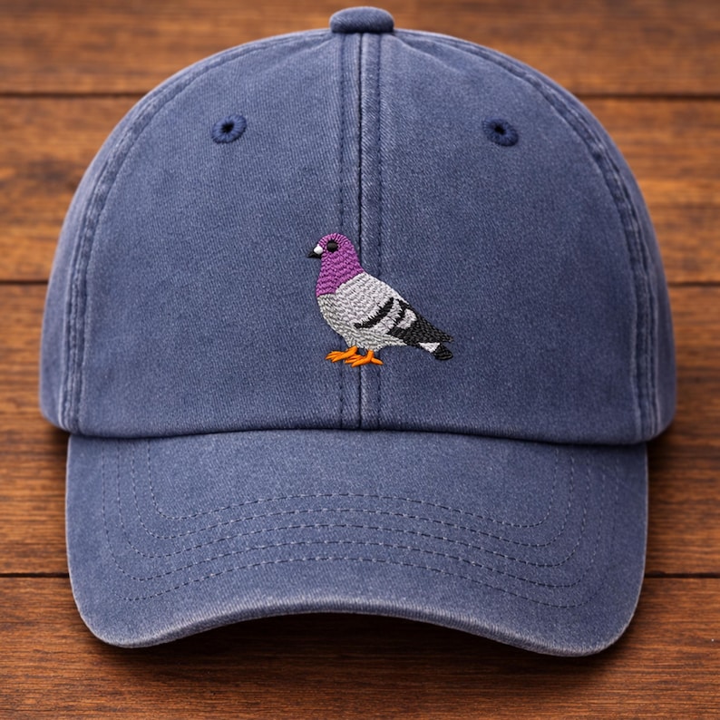 May include: A blue baseball cap with a curved brim. The cap features an embroidered pigeon design with purple, gray, black, and orange accents. The cap has a button on top and a strap in the back.