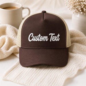 Può includere: Un cappellino trucker marrone con un pannello frontale rigido caratterizzato dalla scritta ricamata in bianco Custom Text, una visiera curva e un pannello posteriore in rete beige con chiusura a scatto regolabile e corona strutturata.
