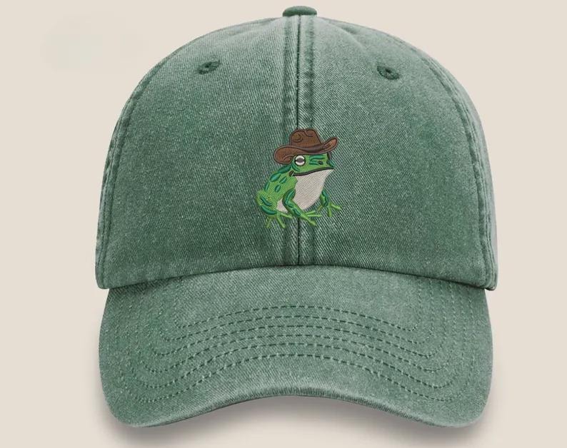 May include: A green baseball cap with a curved peak. The cap has an embroidered design of a frog wearing a brown cowboy hat. The frog is green and white. The cap is made of a soft, worn material.