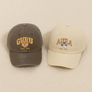 May include: Two baseball caps. One is a dark grey with the word "GURU" in gold lettering and a cat graphic. The other is cream-coloured with "AURA" in brown lettering and a cat graphic. Both caps have curved brims.