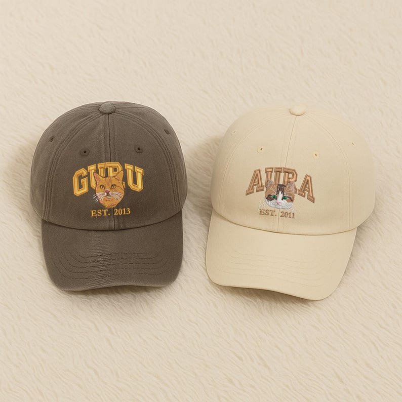 May include: Two baseball caps. One is a dark grey with the word "GURU" in gold lettering and a cat graphic. The other is cream-coloured with "AURA" in brown lettering and a cat graphic. Both caps have curved brims.