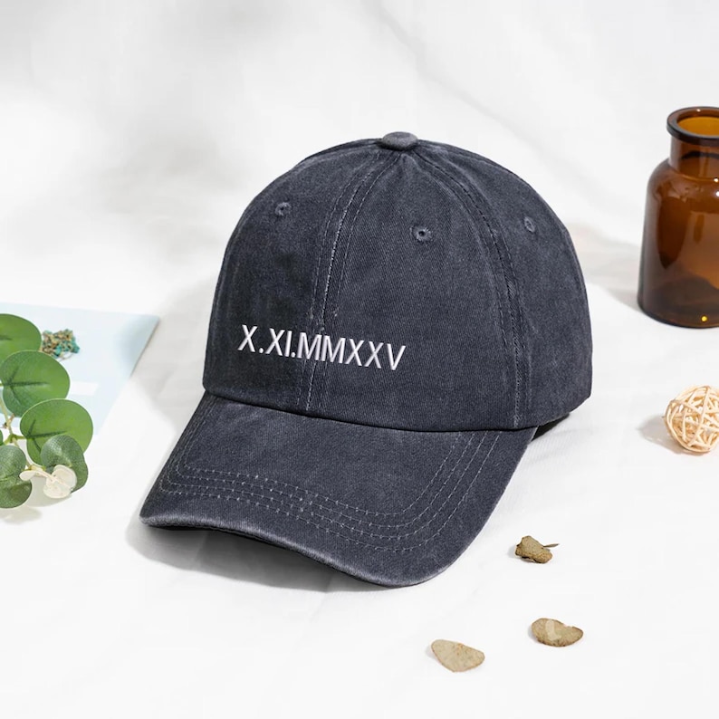 May include: A dark gray baseball cap with a curved brim. The cap features the Roman numeral text "X.XI.MMXXV" in white on the front. The hat is made of a soft fabric and has a classic six-panel design.