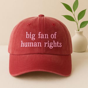 May include: A red baseball cap with the embroidered text "big fan of human rights" in pink. The cap has a curved peak and a fabric strap closure. The hat is a casual accessory.