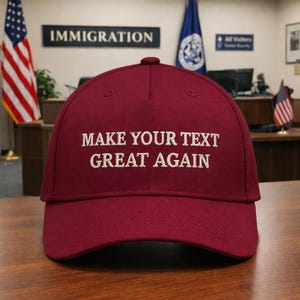 May include: A burgundy baseball cap with the embroidered text "MAKE YOUR TEXT GREAT AGAIN" in white. The cap is sitting on a wooden surface, with flags and a sign that says "IMMIGRATION" in the background.