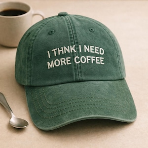 May include: Green baseball cap with the embroidered text "I THINK I NEED MORE COFFEE" in white. The cap has a curved peak and a button on top. A coffee mug and spoon are in the background.