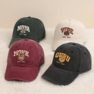 May include: Four baseball caps in different colours: dark green, white, burgundy, and charcoal. Each cap has embroidered text and a cat graphic. The text includes the words "NOVA", "VIBE", "MONK", and "GURU", along with the year of establishment.