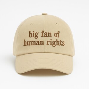 May include: A beige baseball cap with the embroidered text "big fan of human rights" in brown. The cap has a curved brim and a button on top. The hat is a neutral color and suitable for everyday wear.
