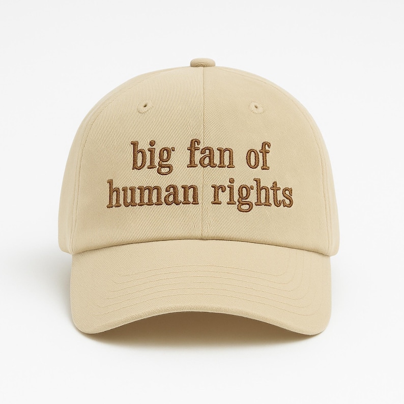 May include: A beige baseball cap with the embroidered text "big fan of human rights" in brown. The cap has a curved brim and a button on top. The hat is a neutral color and suitable for everyday wear.