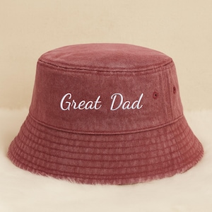 May include: A burgundy bucket hat with the embroidered text "Great Dad" in white. The hat has a classic, rounded shape with a slightly sloped brim. The fabric appears to be a soft, washed cotton or similar material.