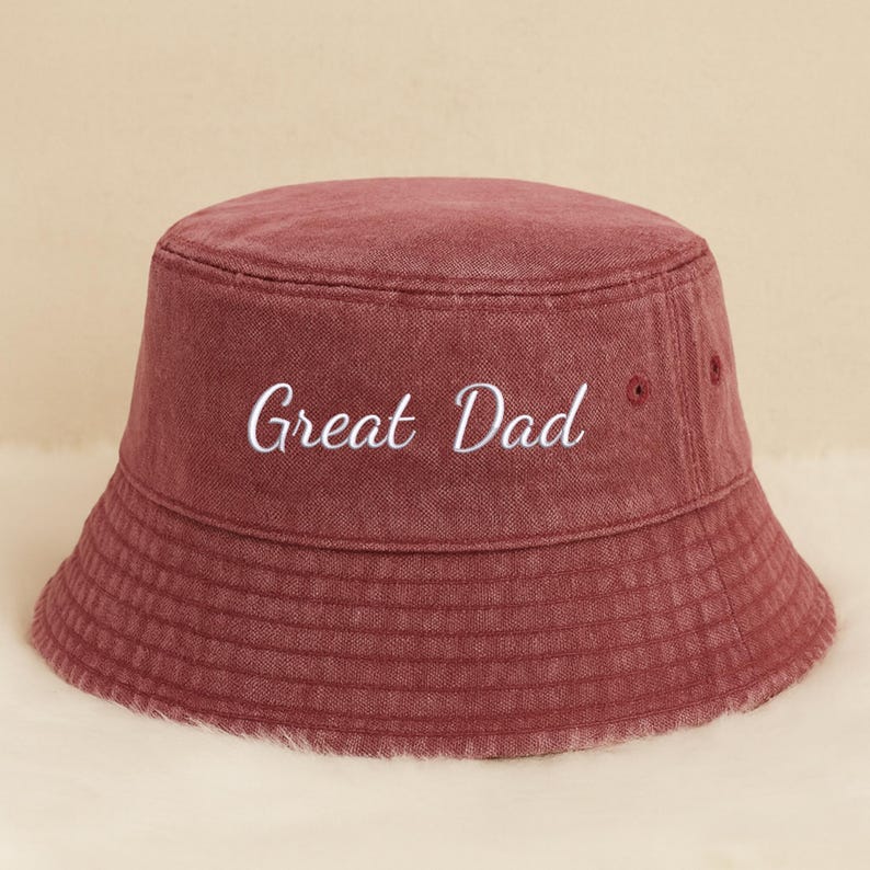 May include: A burgundy bucket hat with the embroidered text "Great Dad" in white. The hat has a classic, rounded shape with a slightly sloped brim. The fabric appears to be a soft, washed cotton or similar material.