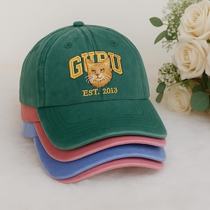 May include: A stack of baseball caps in green, pink, and blue. The top cap is green with the word "GURU" embroidered in gold, a cat face, and the text "EST. 2013". The caps are made of a soft fabric.