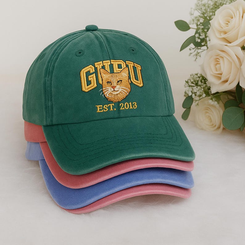May include: A stack of baseball caps in green, pink, and blue. The top cap is green with the word "GURU" embroidered in gold, a cat face, and the text "EST. 2013". The caps are made of a soft fabric.