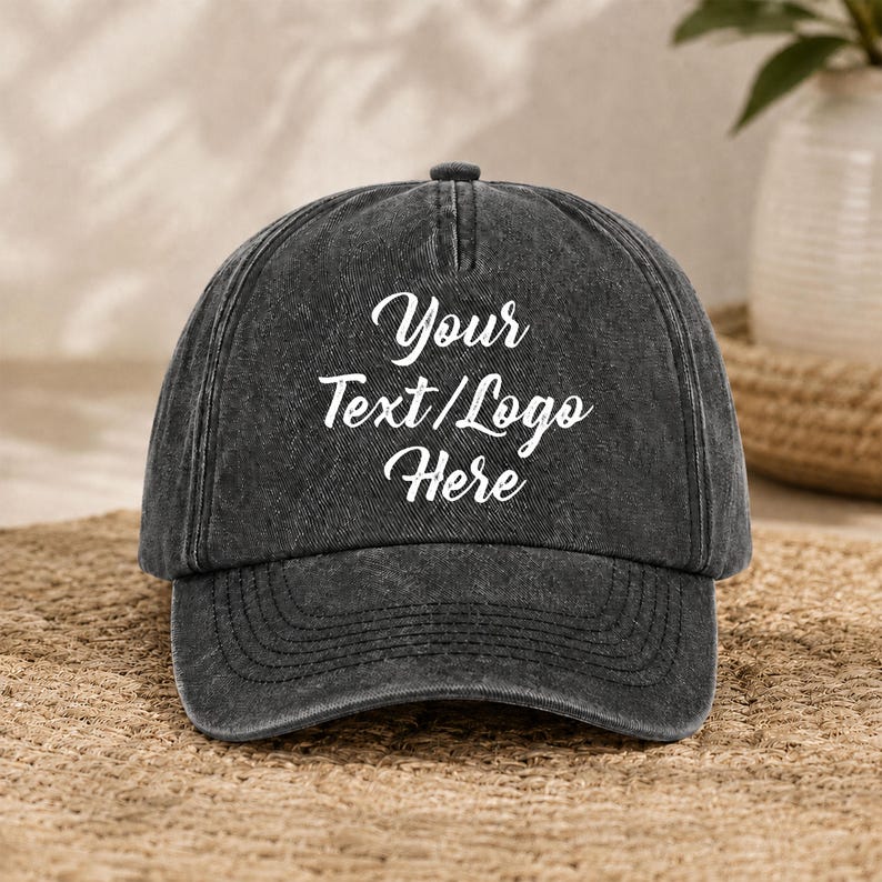 Puede incluir: Una gorra de b&eacute;isbol gris carb&oacute;n con un acabado desgastado y lavado. El panel frontal presenta un texto blanco en cursiva que dice Your Text/Logo Here. La gorra tiene una corona estructurada, una visera curva y costuras visibles.