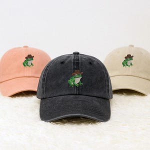 May include: Three baseball caps in peach, charcoal grey, and beige. Each cap has an embroidered green frog wearing a brown cowboy hat. The caps feature a curved brim and an adjustable strap at the back.