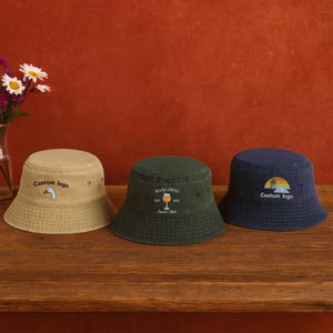 May include: Three bucket hats in tan, dark green, and navy blue sit on a wooden surface. The tan hat has a custom logo of a goose, the green hat says "Margarita Est 2023" and the blue hat has a sunset design with a custom logo.