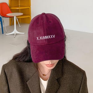 May include: A burgundy baseball cap with the text "X.XI.MMXXV" embroidered in white. The cap is made of a soft material and has a curved brim. The cap is a deep red color.