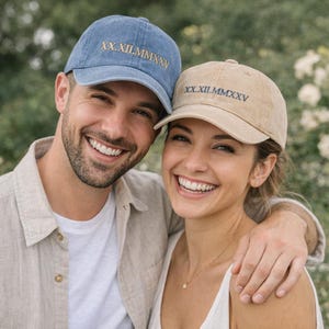 May include: Two baseball caps, one blue and one tan, are shown. Both caps have the Roman numeral date "XX.XII.MMXXV" embroidered on the front. The caps are worn by two people smiling at the camera.