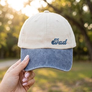 Op de afbeelding: Een tweekleurige baseballpet met een crèmekleurige kroon en een blauwe klep. Het woord "Dad" is in een scriptletter op de voorkant van de pet geborduurd. De pet wordt door een persoon vastgehouden.