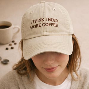 May include: A beige baseball cap with the text "I THINK I NEED MORE COFFEE" in brown lettering. The cap is worn by a person, with a coffee cup and coffee beans in the background.