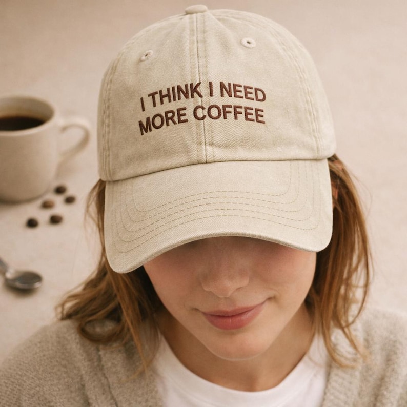 May include: A beige baseball cap with the text "I THINK I NEED MORE COFFEE" in brown lettering. The cap is worn by a person, with a coffee cup and coffee beans in the background.