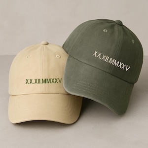 May include: Two baseball caps, one tan and one olive green, are displayed. Each cap has the Roman numeral date "XX.XII.MMXXV" embroidered on the front. The caps are made of a soft fabric and have a classic, curved brim.