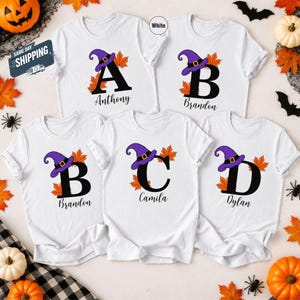 Könnte beinhalten: Fünf weiße kurzärmelige T-Shirts mit großen schwarzen Buchstaben A, B, C und D. Jeder Buchstabe ist mit einem violetten Hexenhut und orangefarbenen Ahornblättern verziert. Die Namen Anthony, Brandon, Camila und Dylan sind darunter gedruckt.
