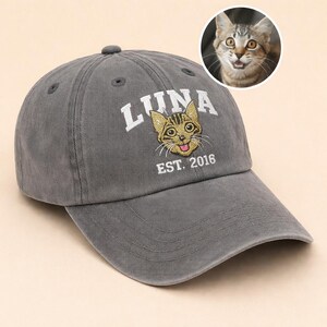 May include: A grey baseball cap featuring the name "LUNA" in white embroidery, a cat graphic, and "EST. 2016". A circular image of a cat is in the upper right. This cap is a casual accessory.