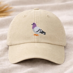 May include: A beige baseball cap featuring an embroidered pigeon design. The bird has a purple head, gray body, black and white wings, and orange feet. The cap has a curved brim and an adjustable strap.