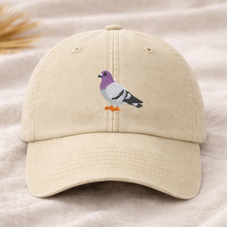 May include: A beige baseball cap featuring an embroidered pigeon design. The bird has a purple head, gray body, black and white wings, and orange feet. The cap has a curved brim and an adjustable strap.