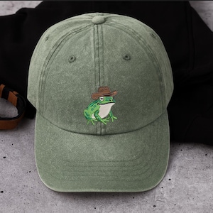 May include: A sage green baseball cap with an embroidered cartoon frog wearing a brown cowboy hat. The frog is green with white details. The cap has a curved peak and an adjustable strap.