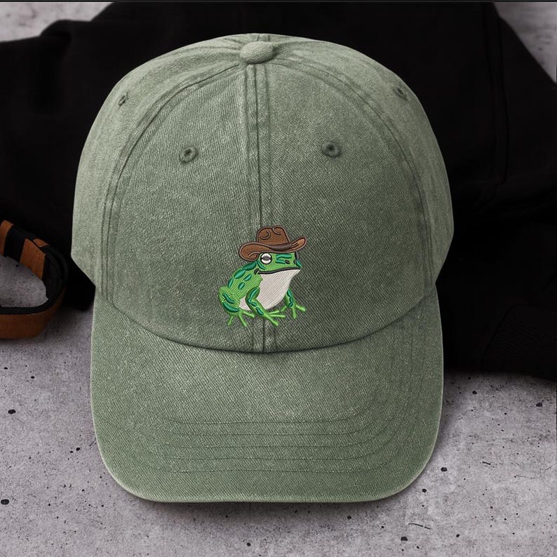 May include: A sage green baseball cap with an embroidered cartoon frog wearing a brown cowboy hat. The frog is green with white details. The cap has a curved peak and an adjustable strap.