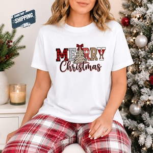 Może przedstawiać: Biała koszulka z krótkim rękawem z napisem Merry Christmas ozdobną czcionką. Litery wypełnione są czerwono-czarną kratą, wzorem w panterkę i srebrnym brokatem. Pośrodku znajduje się choinka w panterkę z czerwoną kokardą.