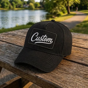May include: Black baseball cap with a curved peak and a rectangular patch. The patch displays the word "Custom" in white script. The cap is on a wooden picnic table near a lake.