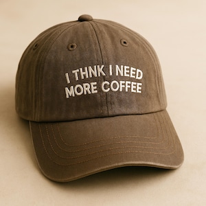 May include: A brown baseball cap with the embroidered text "I THNK I NEED MORE COFFEE" in white. The cap has a curved peak and a button on top. The hat is made of a soft, durable material.
