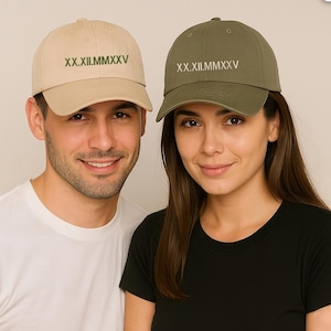 May include: Two baseball caps, one tan and one olive green, each with the Roman numeral text "XX.XII.MMXXV" embroidered on the front. The tan cap is worn by a person wearing a white t-shirt, and the olive green cap is worn by a person wearing a black t-shirt.