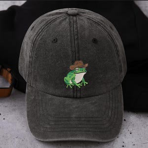 May include: A dark grey baseball cap with a curved peak. The cap has an embroidered green frog wearing a brown cowboy hat. The frog has white detailing and is centered on the front. The cap is made of a textured fabric.